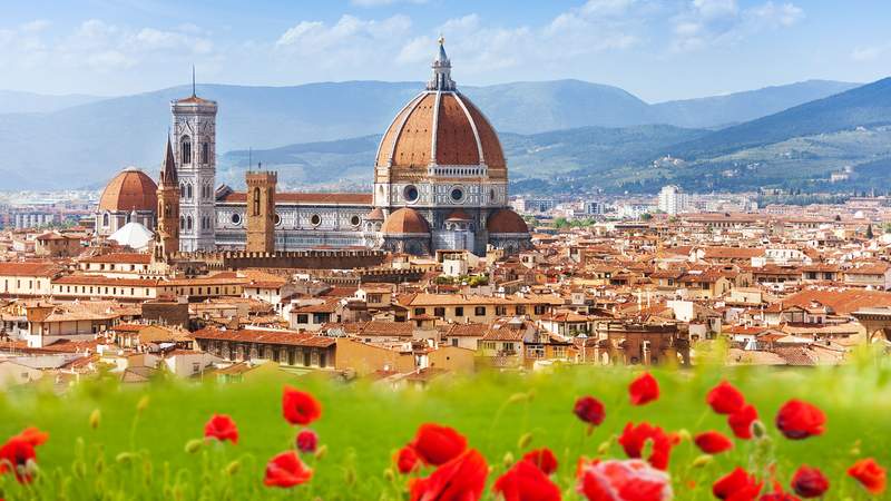 Man sieht die Stadt Florenz von schräg oben. Besonders der Dom ist gut zu sehen. Im Vordergrund sind rote Mohnblumen zu sehen und im Hintergrund die Berge