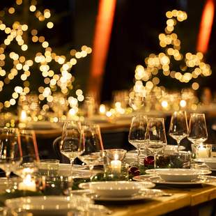 Weihnachtsfeier "Herzhaft & Festlich" Festlich gedeckte Tafel für eine Weihnachtsfeier mit elegant arrangierten Gläsern und Tellern, umgeben von warmem Kerzenschein und verschwommenen, leuchtenden Weihnachtslichtern im Hintergrund in Form von Sternen und Bäumen.