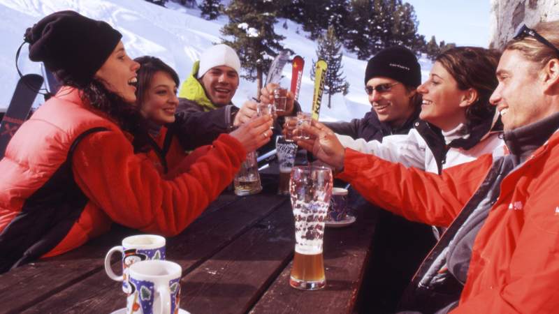 6 Personen sitzen im Außenbereich einer Hütte im Winter und stoßen mit Getränken an.