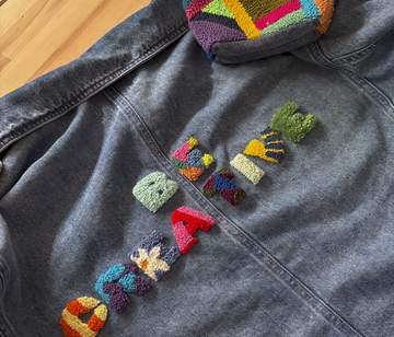 Schriftzug mit Punch Needling auf einer Jeansjacke Jeansjacke auf Holzhintergrund mit bunten Punch-Needle-Buchstaben und geometrischen Patches